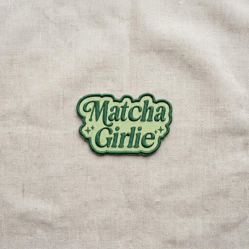 K&ouml;nnte beinhalten: Ein gr&uuml;n-hellgr&uuml;ner, gestickter Aufn&auml;her mit dem Schriftzug "Matcha Girlie" in einer verspielten Schriftart. Der Aufn&auml;her hat einen hellgr&uuml;nen Hintergrund und einen dunkelgr&uuml;nen Rand. Auf beiden Seiten des Textes befinden sich zwei kleine sternf&ouml;rmige Designs.