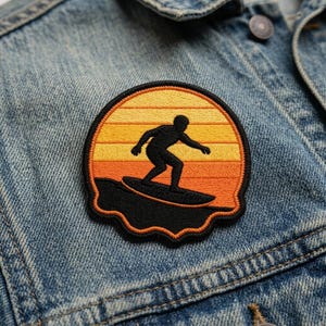 May include: Embroidered patch featuring a surfer silhouetted against a sunset. The patch has a black border and a design of orange, yellow, and black. The patch is attached to a denim jacket.