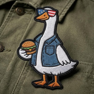 May include: Embroidered patch featuring a white goose wearing a denim jacket and sunglasses with an American flag design. The goose is holding a hamburger. The patch has a black border and is set against an olive green background.