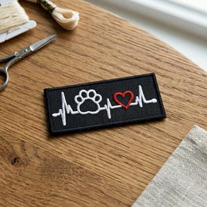 May include: Black rectangular patch with a white paw print, a red heart, and a white heartbeat line design. The patch is embroidered and likely intended for clothing or accessories. The design suggests a pet-themed item.
