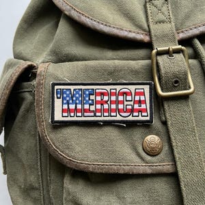 May include: An olive green canvas backpack with brown leather trim and a rectangular patch that reads "'MERICA" in red, white, and blue. The backpack has a flap pocket, a buckle closure, and a brass button.