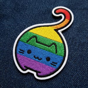 May include: Embroidered patch featuring a cartoon cat in a rainbow design. The cat has a white outline and a curved tail. The rainbow colors include red, orange, yellow, green, blue, and purple. The patch is on a blue denim background.