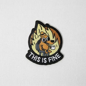 May include: Embroidered patch featuring a cartoon dog sitting in a chair, surrounded by flames, holding a coffee cup. The text "THIS IS FINE" is below the dog in a black banner. The dog is brown, the flames are yellow and orange, and the text is white.