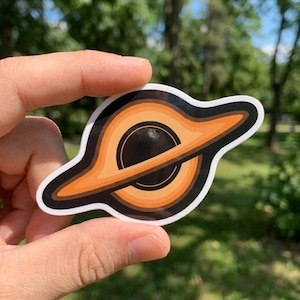 May include: A die-cut sticker featuring a black hole illustration with a central dark sphere surrounded by a bright orange and light orange accretion disk, all outlined in a thick white border against a blurred natural outdoor background.