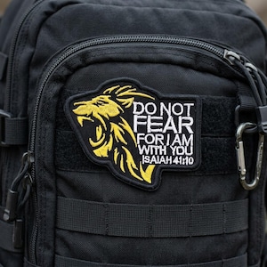 May include: Black tactical backpack with a yellow and black patch. The patch features a roaring lion and the text "DO NOT FEAR FOR I AM WITH YOU ISAIAH 41:10". The backpack has multiple straps and a metal carabiner.