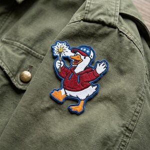 May include: Embroidered patch featuring a cartoon goose wearing a red hoodie and a blue and white hat, holding a sparkler. The patch has a blue border and is affixed to an olive green jacket.