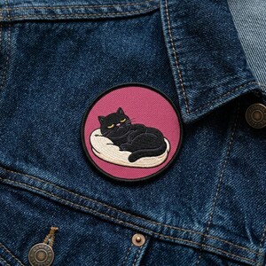 May include: Embroidered patch featuring a black cat resting on a white pillow, set against a pink background. The cat has yellow eyes and is lying down. The patch is circular and is attached to a denim jacket.