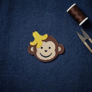 May include: Embroidered patch featuring a smiling monkey with a yellow banana on its head. The monkey has a brown face and ears, with a tan face and a black stitched smile. A spool of brown thread and small scissors are in the background.