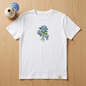 May include: White t-shirt featuring a detailed illustration of blue hydrangea flowers tied with a blue ribbon. The floral design is centered on the chest. A small, off-white tag with text is sewn on the bottom right.
