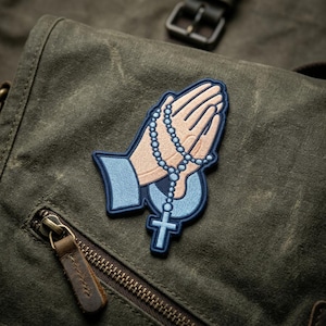 May include: Embroidered patch featuring praying hands holding a rosary with a cross pendant. The patch has a light blue border and accents, with a beige and light blue color scheme. The patch is placed on a textured, olive green fabric.