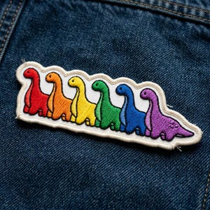 May include: Embroidered patch featuring six colorful dinosaurs in a rainbow spectrum. Each dinosaur is a different color, from red to purple, with a white border. The patch is displayed on a denim background.