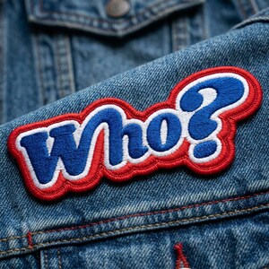May include: Embroidered patch with the word "Who?" in blue, white, and red. The patch is sewn onto a denim jacket. The text is in a bold, playful font, with a question mark.