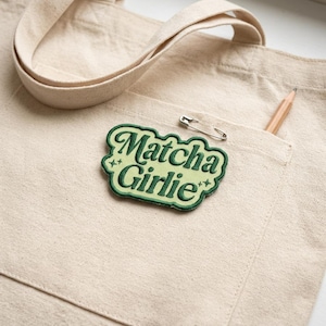 May include: A beige canvas tote bag with a pocket, featuring a green and white embroidered patch that reads "Matcha Girlie." A silver safety pin and a wooden pencil are also in the pocket. The bag's strap is visible.