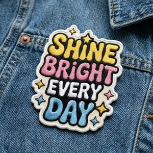 May include: Embroidered patch with the words "Shine Bright Every Day" in yellow, pink, white, and blue. The patch is outlined in white and features star accents. It is affixed to a denim jacket.