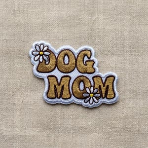 May include: Embroidered patch with the words "DOG MOM" in brown, outlined in white and light blue. The patch features daisy accents. The patch is on a beige fabric background.