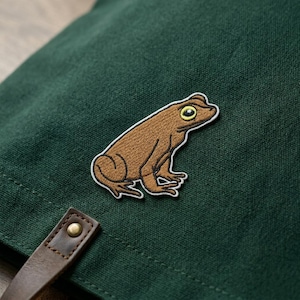 May include: A brown frog patch with a white border is sewn onto a dark green fabric. The frog has yellow eyes and is in a sitting position. A brown leather strap with a brass rivet is visible in the lower left corner.