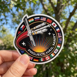 May include: A circular sticker featuring a central illustration of a white spacecraft with a red curved trail against a black background. The text PROJECT HAIL MARY is printed in white along the bottom. A border of various national flags surrounds the center.