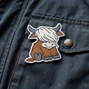 May include: Embroidered patch featuring a Highland cow design. The patch has a brown body, white shaggy fur, and gray horns. It is sewn onto a dark blue jacket near a pocket with a brass button.