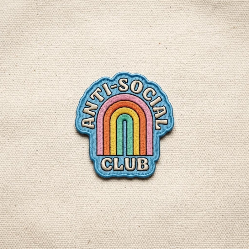 以下が含まれることがあります： 「ANTI-SOCIAL CLUB」の文字が虹のデザインを囲む青いパッチ。虹はオレンジ、黄色、緑、ピンク、赤のアーチが特徴です。パッチはクリーム色の生地の上にあります。