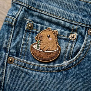 May include: Embroidered patch featuring a cartoon capybara enjoying a drink in a coconut shell. The patch is brown, tan, and white, with a green and white straw. The patch is attached to a blue denim pocket.