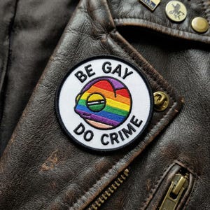 May include: A round, embroidered patch with the text "BE GAY DO CRIME" encircling a rainbow-colored chameleon design. The patch is affixed to a brown leather jacket, with visible zippers and metal pins.