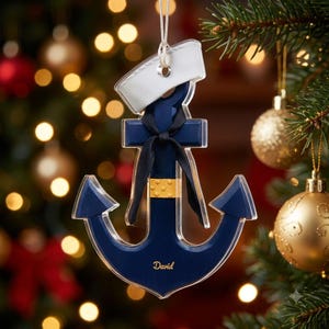 May include: A navy blue anchor-shaped Christmas ornament with a clear acrylic border. It features a white sailor hat on top, a black ribbon tied in a bow, a gold band with dots, and the name David printed in gold lettering on the bottom.