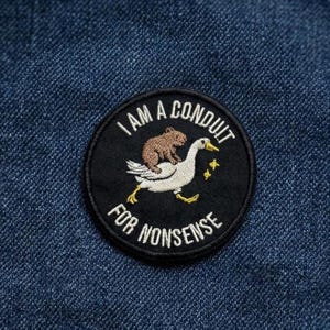 May include: A round black patch with the text "I AM A CONDUIT FOR NONSENSE". The patch features an illustration of a brown bear riding on the back of a white goose, with yellow stars.