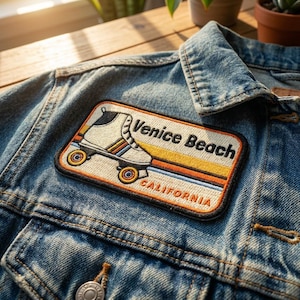 May include: A blue denim jacket featuring a rectangular embroidered patch. The patch displays a white roller skate with orange and blue stripes, the text Venice Beach in black, and the word CALIFORNIA in orange against a sunset-colored background.