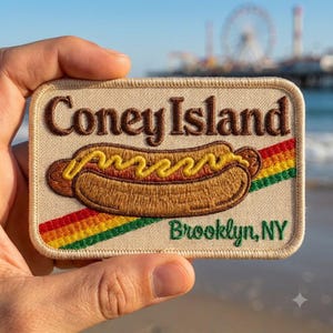 May include: A rectangular embroidered patch with a beige background featuring the brown text Coney Island and green text Brooklyn, NY. A brown hot dog with yellow mustard is centered, accented by a diagonal red, yellow, and green stripe.