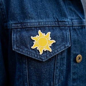 May include: A yellow sun-shaped enamel pin with a white outline is affixed to the pocket of a blue denim jacket. The pin features a bright yellow sun with detailed rays and a circular center.