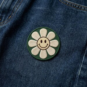 May include: Embroidered patch featuring a smiling daisy design. The daisy has white petals, a yellow center, and a green border. The patch is affixed to a blue denim fabric, showcasing a casual, cheerful aesthetic.