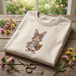 Op de afbeelding: Een beige sweatshirt met een geborduurde konijn met een bloemenkrans en een patchwork jurk, met een mandje wortels. Gouden schaar en bloemen zijn gerangschikt op een houten oppervlak, wat een lente- of Paas thema creëert.