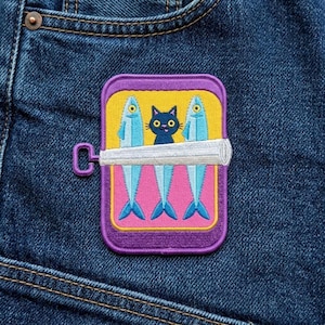 May include: A purple, yellow, and pink embroidered patch featuring a sardine can design. The can contains two blue fish and a black cat. The patch is attached to a blue denim fabric.