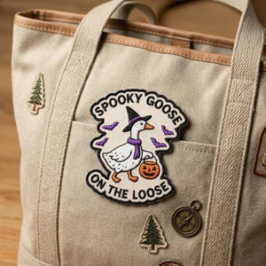 May include: Beige canvas tote bag with a "Spooky Goose on the Loose" patch. The patch features a goose wearing a witch hat and holding a jack-o'-lantern. Additional patches include a pine tree and a compass.