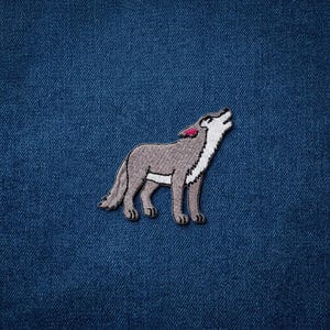 May include: Embroidered patch featuring a howling wolf in shades of gray, white, and pink. The wolf is depicted in a side profile, with its head raised and mouth open. The patch is set against a textured blue background.