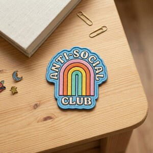 以下が含まれることがあります： 「ANTI-SOCIAL CLUB」の文字が虹のデザインを囲む青いパッチ。虹はピンク、オレンジ、黄色、緑、青のアーチが特徴です。パッチは、本、ペーパークリップ、小さなピンが置かれた木製の表面にあります。