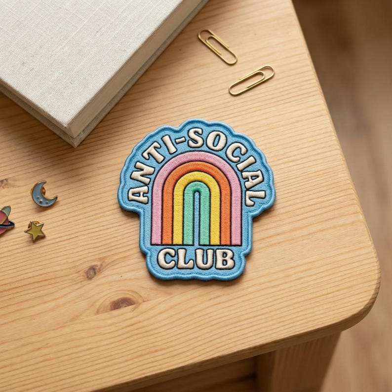 以下が含まれることがあります： 「ANTI-SOCIAL CLUB」の文字が虹のデザインを囲む青いパッチ。虹はピンク、オレンジ、黄色、緑、青のアーチが特徴です。パッチは、本、ペーパークリップ、小さなピンが置かれた木製の表面にあります。