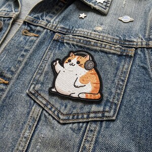 May include: Embroidered patch featuring a cartoon cat wearing headphones and giving a thumbs up. The cat has white and orange fur and is outlined in black. The patch is on a denim jacket.