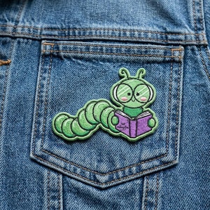 May include: A green caterpillar patch with glasses, reading a purple book, is sewn onto a denim jacket pocket. The caterpillar has a smiling face and is surrounded by a green outline. The book has the text "middaylos" on the cover.