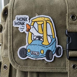 May include: Embroidered patch featuring a goose driving a blue toy car. The patch has the words "HONK HONK" and "SILLY" embroidered on it. The goose has an orange beak. The patch is attached to a green canvas bag.