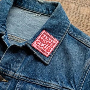 May include: A close-up of a blue denim jacket with a pink patch. The patch, with a red border, reads "BADASS MOMS CLUB" in red lettering. The jacket's collar and a portion of the front are visible, showing the texture of the denim.