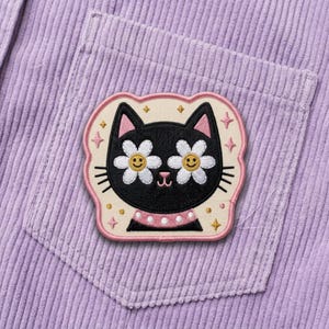 May include: Embroidered patch featuring a black cat with daisy eyes and a pink collar. The patch is set against a light beige background with a pink border and star accents. The patch is sewn onto a lavender corduroy pocket.