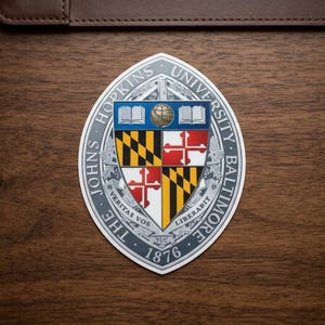 May include: An oval Johns Hopkins University sticker with a detailed emblem. The emblem features a shield with a globe, books, and a cross design, surrounded by text. The sticker is on a wooden surface.