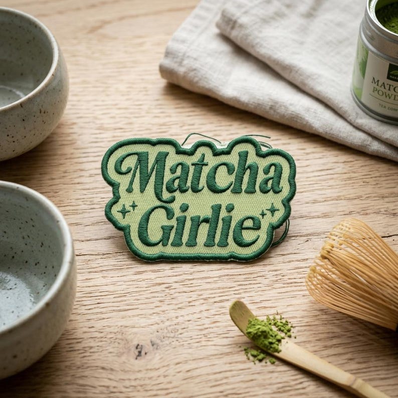 K&ouml;nnte beinhalten: Ein gr&uuml;ner, gestickter Aufn&auml;her mit den Worten "Matcha Girlie" in einer stilisierten Schriftart. Der Aufn&auml;her liegt auf einer Holzoberfl&auml;che, zusammen mit Matcha-Pulver, einem Schneebesen und Teeschalen. Der Aufn&auml;her ist ein dekorativer Artikel.