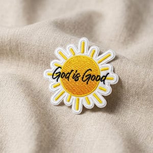 May include: Embroidered patch in the shape of a sun with a yellow center and white and yellow rays. The phrase "God is Good" is written in black script across the sun.