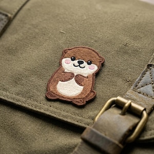 May include: A brown otter patch with a smiling face and pink cheeks is sewn onto a green canvas bag. The otter is holding its paws together. The bag has a leather strap and a metal buckle.