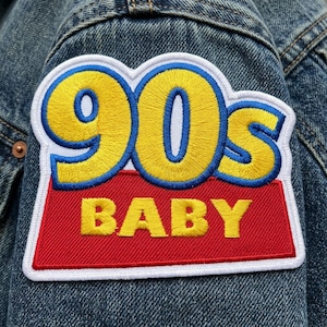 May include: Embroidered patch with the text "90s BABY" in yellow, blue, and white on a red background. The patch is sewn onto a denim jacket. The patch is rectangular with rounded corners.
