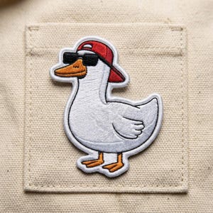 May include: Embroidered patch featuring a white goose wearing a red baseball cap and black sunglasses. The goose has an orange beak and feet. The patch is sewn onto a beige fabric background.