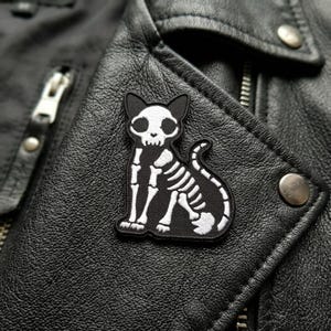 May include: A black and white embroidered patch featuring a cat skeleton design. The patch is affixed to a black leather jacket. The cat skeleton has a skull-shaped head and a detailed rib cage. The patch is a unique accessory.