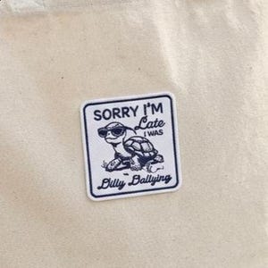 May include: A square patch with a white background and a navy blue border. The patch features a cartoon turtle wearing sunglasses, with the text "SORRY I'M Late (I WAS)" above it, and "Billy Ballying" below.
